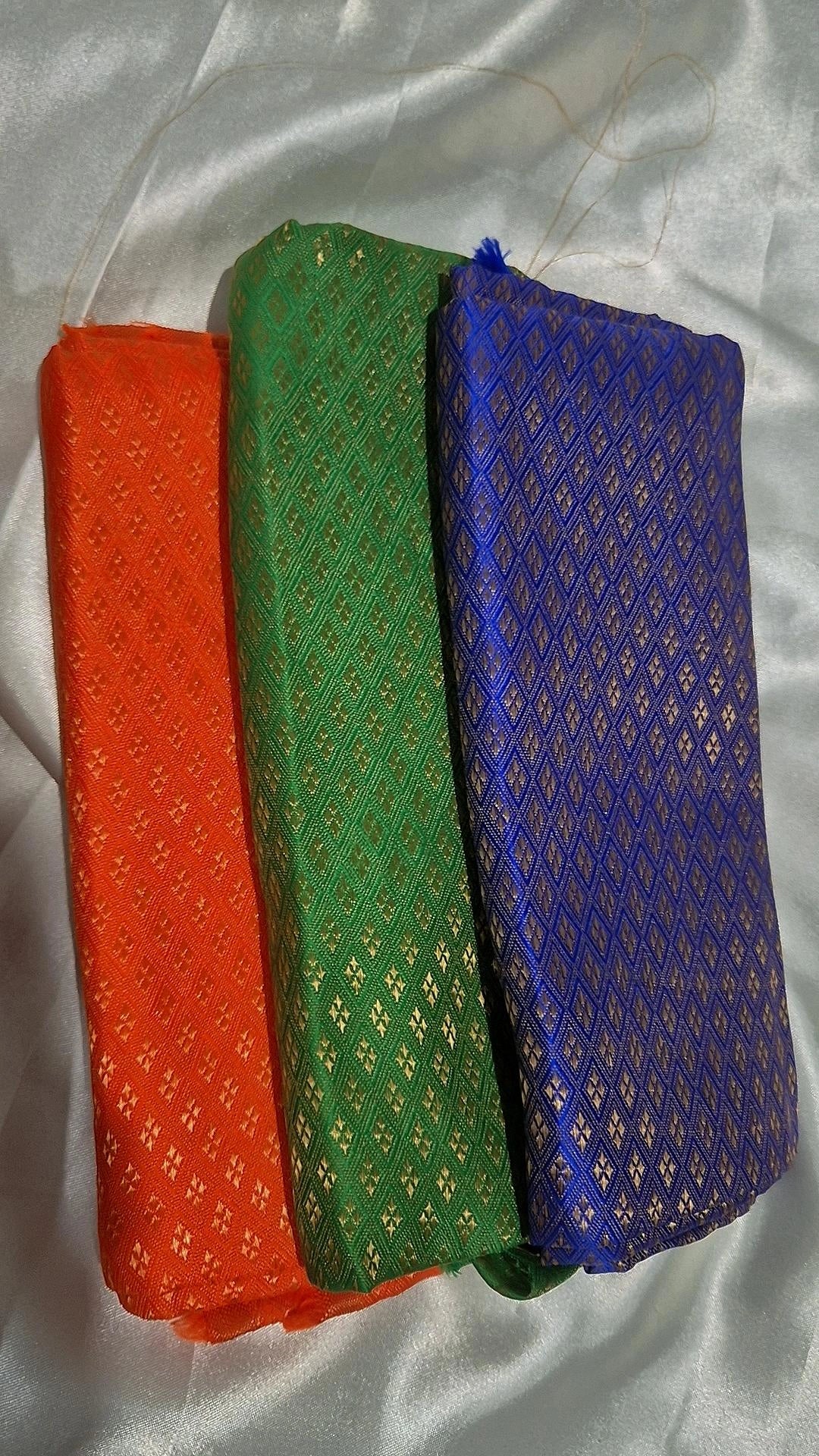 Three Banarasi silk blouse pieces in orange, green and blue folded on white satin