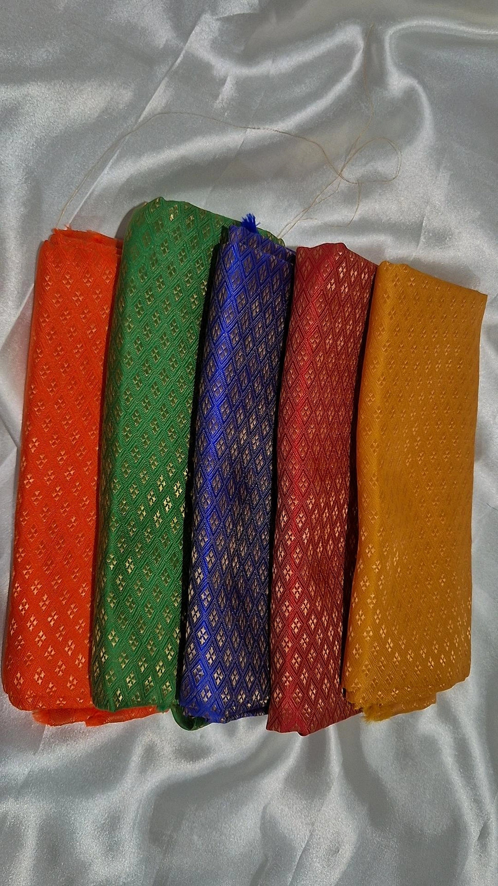 Five Banarasi silk blouse pieces in orange, green, blue, red and mustard fanned out on white satin