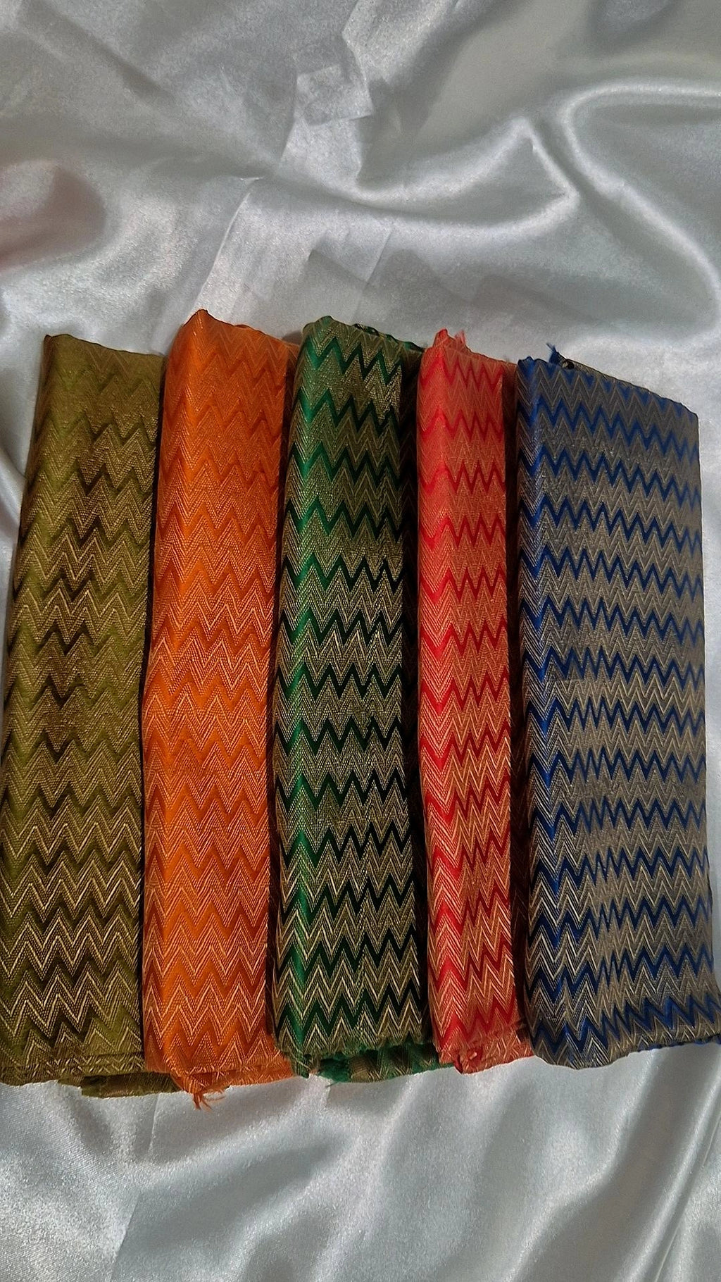 Five Banarasi silk chevron weave blouse pieces in olive, orange, green, red and blue fanned out on white satin