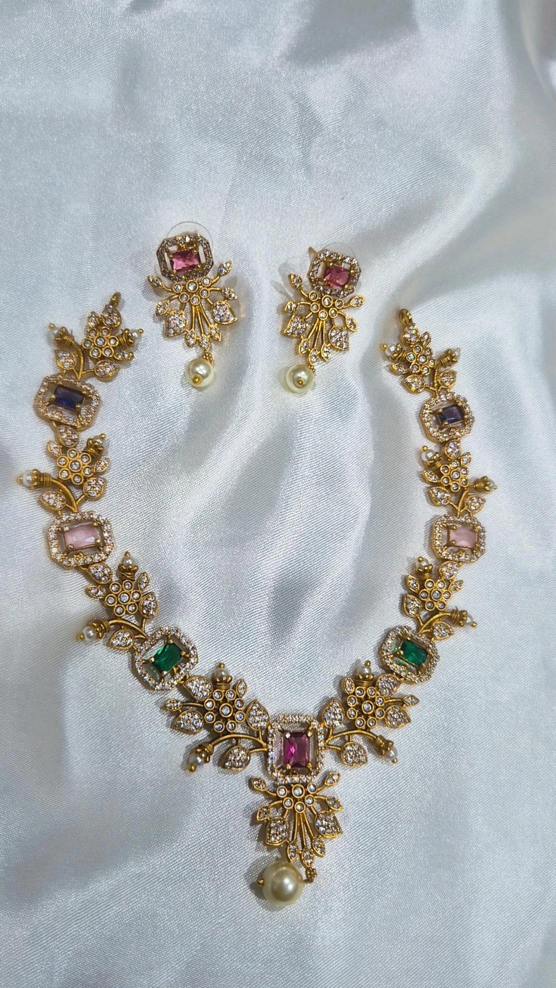 Multicolour CZ floral necklace set with ruby, amethyst, blush pink and emerald rectangular stones, white CZ pave links and pearl drop pendant with matching earrings on white satin