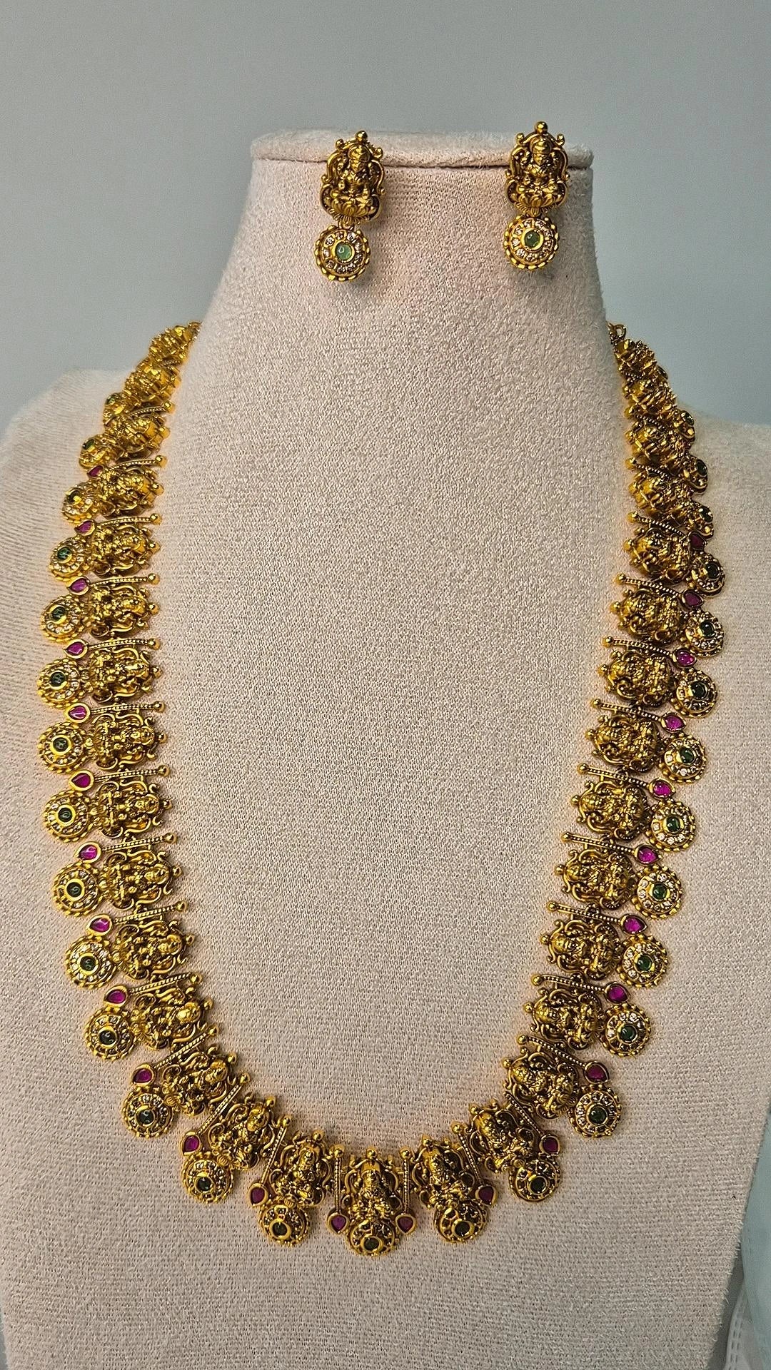 Lakshmi temple gold long haram necklace with intricate carved motifs and ruby stone accents displayed on jewellery bust with earrings